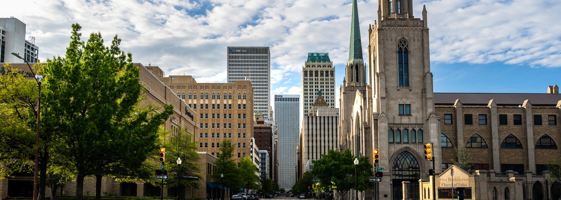 Downtown Tulsa street scene: First Presbyterian Church of Tulsa's stone tower and green copper steeple at the right, downtown towers including BOK Tower in the distance, and leafy green trees lining the street